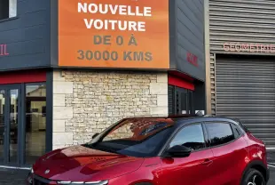 Vue 3/4 avant droite d'une Alfa Romeo Junior rouge avec toit noir, jantes noires et calandre distinctive devant un bâtiment commercial.