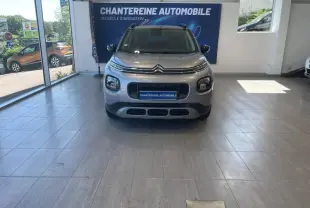 Vue frontale d'un Citroën C3 Aircross gris acier exposé en showroom avec barres de toit noires visibles.