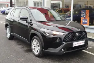 Toyota Corolla Cross noir intense vue 3/4 avant droit, avec calandre noire et feux LED allumés, stationnée devant un showroom.