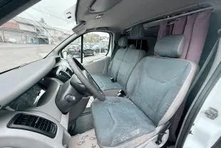 Vue intérieure côté conducteur du Nissan Primastar blanc de 2005, montrant les sièges en tissu gris bleu et le tableau de bord.