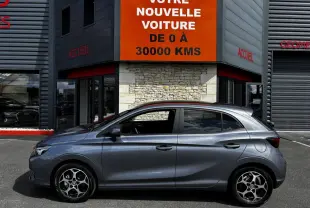Vue latérale gauche d'une MG3 gris clair 2025 garée devant un bâtiment avec panneau orange "Votre nouvelle voiture".