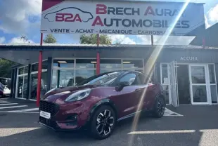 Ford Puma rouge Vivid Ruby en 3/4 avant droit devant un garage avec enseigne Brech Aura Automobiles