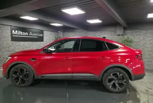 Profil droit du Renault Arkana 2023 rouge flamme avec jantes noires et toit ouvrant électrique visible en intérieur showroom.