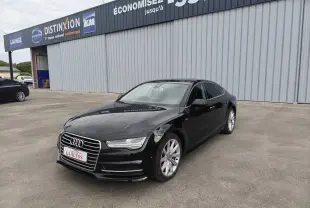 Audi A7 noire vue en 3/4 avant droit, avec jantes aluminium et calandre distinctive, stationnée devant un bâtiment commercial.
