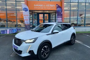 Peugeot 2008 blanc vue 3/4 avant droit, jantes alliage bi-ton et calandre noire devant un garage.