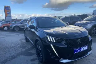 Peugeot 2008 noir vue 3/4 avant droit, avec calandre distinctive et feux LED verticaux allumés sur parking extérieur.