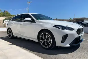 BMW Série 1 blanche vue 3/4 avant droit, avec jantes alliage bicolores et calandre M Sport sous un ciel bleu.