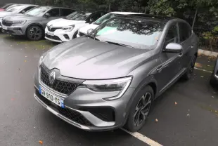 Renault Arkana gris métal foncé vu en 3/4 avant droit sur un parking sous un ciel couvert.