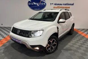Dacia Duster blanc en 3/4 avant droit, avec jantes noires et grises, feux allumés et attelage visible.