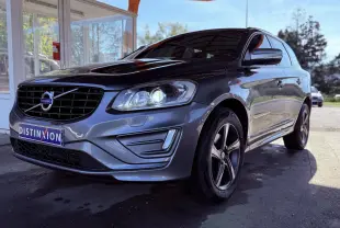 Volvo XC60 gris en 3/4 avant droit, phare allumé et jantes alliage cinq branches visibles, stationné devant un bâtiment.