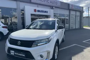 Vue 3/4 avant d'un Suzuki Vitara blanc hybride 2023 garé devant un concessionnaire Suzuki avec logo visible.