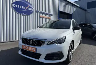 Peugeot 308 SW blanc nacré vue 3/4 avant droit, avec calandre noire et plaque orange "OCCASION" devant un bâtiment industriel.