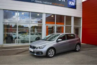 Peugeot 308 gris métallisé vue 3/4 avant droit, stationnée devant une vitrine de concession multimarque.