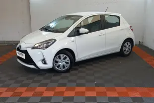 Vue 3/4 avant droit d'une Toyota Yaris blanche hybride 2019 avec jantes acier et feux ellipsoïdaux allumés.