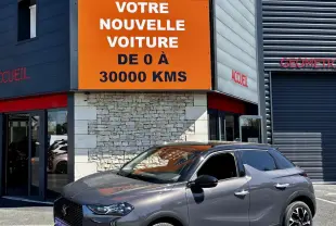 DS3 gris foncé en 3/4 avant droit, stationnée devant un bâtiment avec panneau orange Distinxion visible.