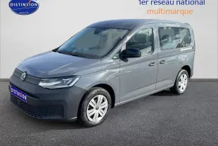 Volkswagen Caddy gris pur en 3/4 avant droit, avec barres de toit et jantes alliage visibles.