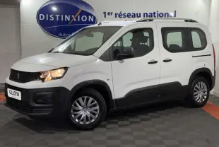 Peugeot Rifter blanc en vue 3/4 avant droit, avec barres de toit chromées et feux allumés dans un showroom.