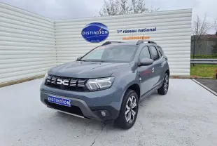 Dacia Duster gris urbain en 3/4 avant droit, avec jantes bi-ton et barres de toit visibles.