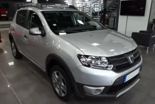 Vue 3/4 avant d'une Dacia Sandero Stepway gris platine en showroom, avec jantes alliage et barres de toit noires.