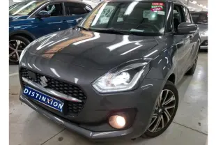 Suzuki Swift grise vue 3/4 avant droit, phares et antibrouillard allumés en intérieur showroom.