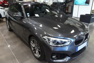 BMW Série 1 F21 LCI gris vue 3/4 avant droit, avec jantes alliage et phares LED allumés en showroom.