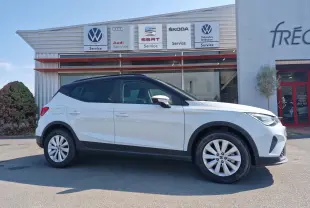 Vue de profil côté gauche d'un SEAT Arona blanc alpin avec toit noir, stationné devant un service multimarques.