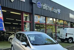 Ford Fiesta gris lunaire métal vue 3/4 avant droit devant un garage avec enseignes commerciales.