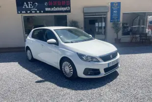 PEUGEOT 308 blanc vue 3/4 avant droit, garée devant un bâtiment avec panneaux publicitaires visibles.
