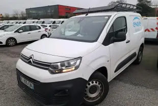 Vue 3/4 avant d’un utilitaire Citroën Berlingo blanc avec pare-chocs noirs, stationné sur un sol gravillonné.