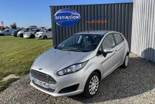 Ford Fiesta 1.25 82 CV Trend gris titane vue 3/4 avant droit sur parking gravillonné devant un mur noir.
