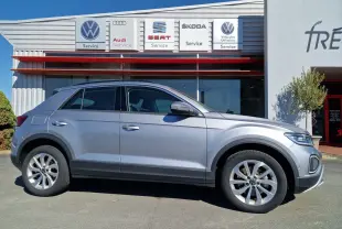 Profil côté gauche d'un Volkswagen T-Roc gris pyrite 2023 garé devant un centre de service multimarques.