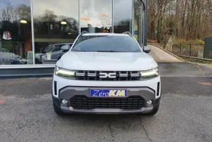 Vue avant d'un Dacia Duster blanc glacier 2025 avec calandre noire et feux LED allumés, stationné devant une vitrine.