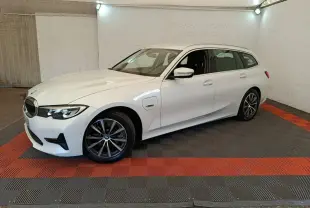 BMW Série 3 320e blanche vue 3/4 avant droit en break, avec jantes alliage et prise de recharge latérale visible.