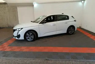 Peugeot 308 blanche vue de profil côté gauche, dans un espace fermé avec sol gris et bandes rouges.