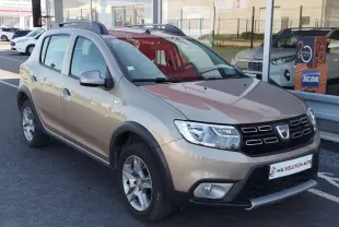 Vue 3/4 avant d'une Dacia Sandero Stepway beige dune avec barres de toit et jantes alliage, stationnée devant un garage.