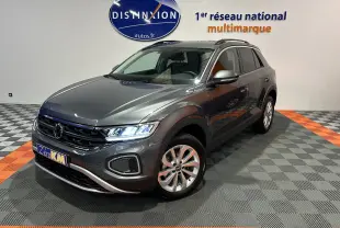 Volkswagen T-Roc gris Indium métal en 3/4 avant droit, avec feux allumés et jantes alliage visibles.