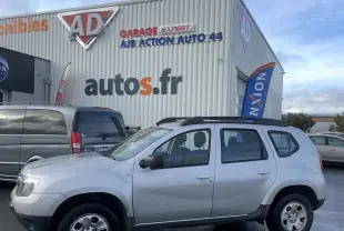 Vue de profil côté gauche d'un Dacia Duster gris avec barres de toit et enjoliveurs, stationné devant un garage.