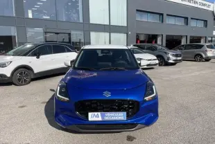 Suzuki Swift 1.2 Dualjet Hybrid bleu métal vue de face, calandre noire et toit blanc visible, devant un garage.