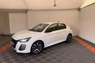Peugeot 208 blanche vue en 3/4 avant droit dans un garage avec jantes noires et phares LED allumés.