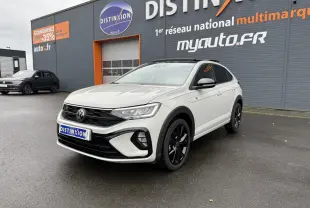 Volkswagen Taigo blanc en 3/4 avant droit avec jantes noires, stationné devant un bâtiment gris et orange.