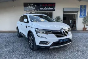 Renault Koleos blanc vu en 3/4 avant droit, avec calandre chromée et jantes alliage, stationné devant un bâtiment.
