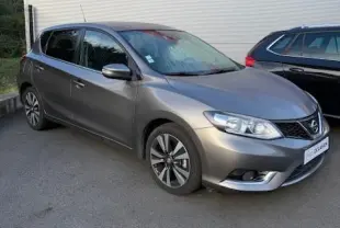 Nissan Pulsar gris foncé vue 3/4 avant droit, avec jantes alliage et calandre chromée, stationnée devant un mur gris.