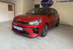 KIA Rio rouge vue 3/4 avant droit en intérieur, avec plaque "OCCASION" et jantes alliage distinctives.