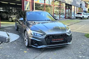 Audi A5 Sportback gris Daytona vue 3/4 avant, avec calandre noire hexagonale et jantes alliage visibles devant un garage.