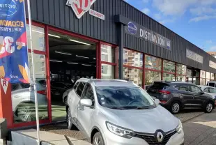 Renault Kadjar gris platine métal en 3/4 avant, stationné devant un garage avec enseignes visibles.
