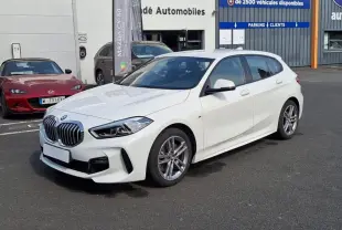 BMW Série 1 120i M Sport blanc vue 3/4 avant droit, avec jantes aluminium et calandre imposante.