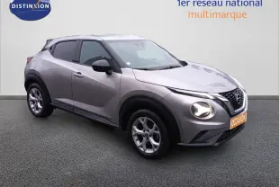 Nissan Juke gris platine métal en 3/4 avant droit, avec phares allumés et jantes alliage visibles.