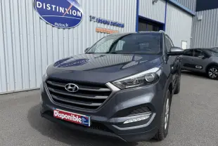 Hyundai Tucson gris en 3/4 avant droit, avec calandre marquée et feux LED, stationné devant un bâtiment commercial.