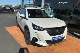 Peugeot 2008 blanc vue 3/4 avant droit, calandre noire et jantes foncées, stationnée devant un showroom.
