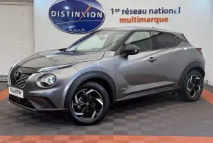 Nissan Juke gris foncé en 3/4 avant droit, avec jantes noires et logo Hybrid visible sur l'aile avant.
